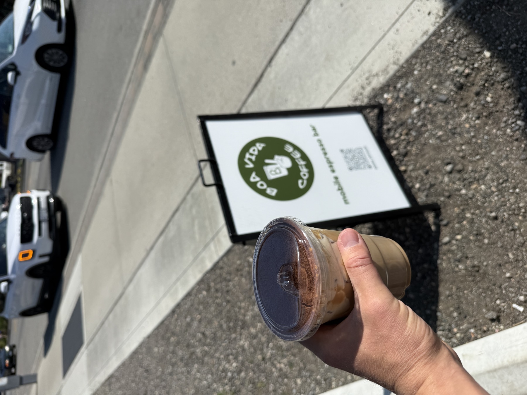 An iced tiramisu latte dusted with cocoa, held in hand beside the Boa Vida Coffee sidewalk sign